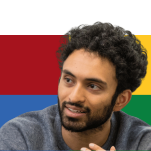 Man with dark curly hair and beard smiling, wearing gray shirt, with colorful red, blue, yellow, and green background blocks