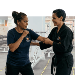 Two women in martial arts attire demonstrate a combat move against a purple wall, representing workplace wellness training