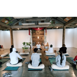 Group yoga class with participants seated on mats in a bright studio with wooden beams and large gold number 8 above