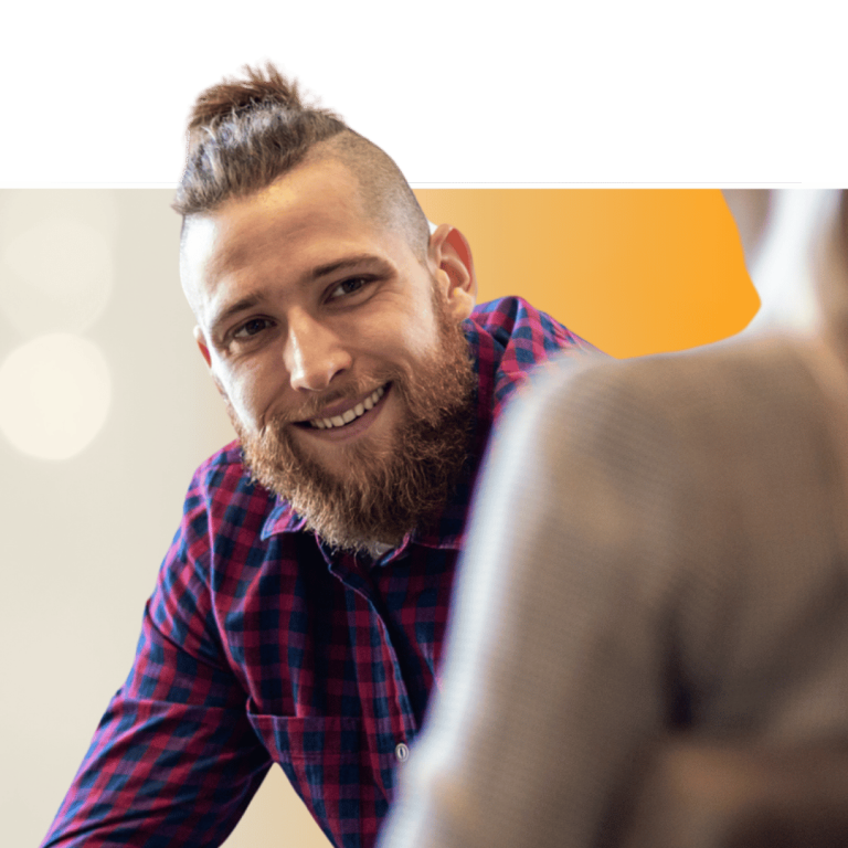Smiling man with beard and mohawk hairstyle in red and blue plaid shirt, engaged in conversation with another person