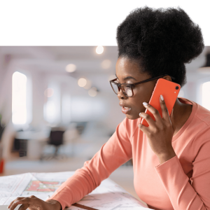 Woman wearing glasses and coral shirt talking on phone while reviewing documents at her desk in a modern office