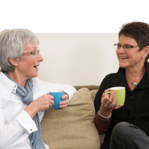 Two women sitting on a couch smiling and holding mugs, representing workplace connection and wellbeing