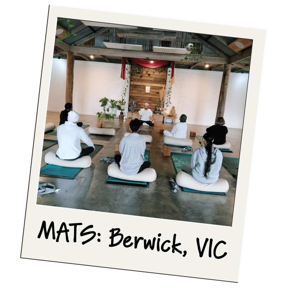 Yoga class in session with instructor leading group of participants seated on mats in a rustic studio space with exposed beams and wooden backdrop